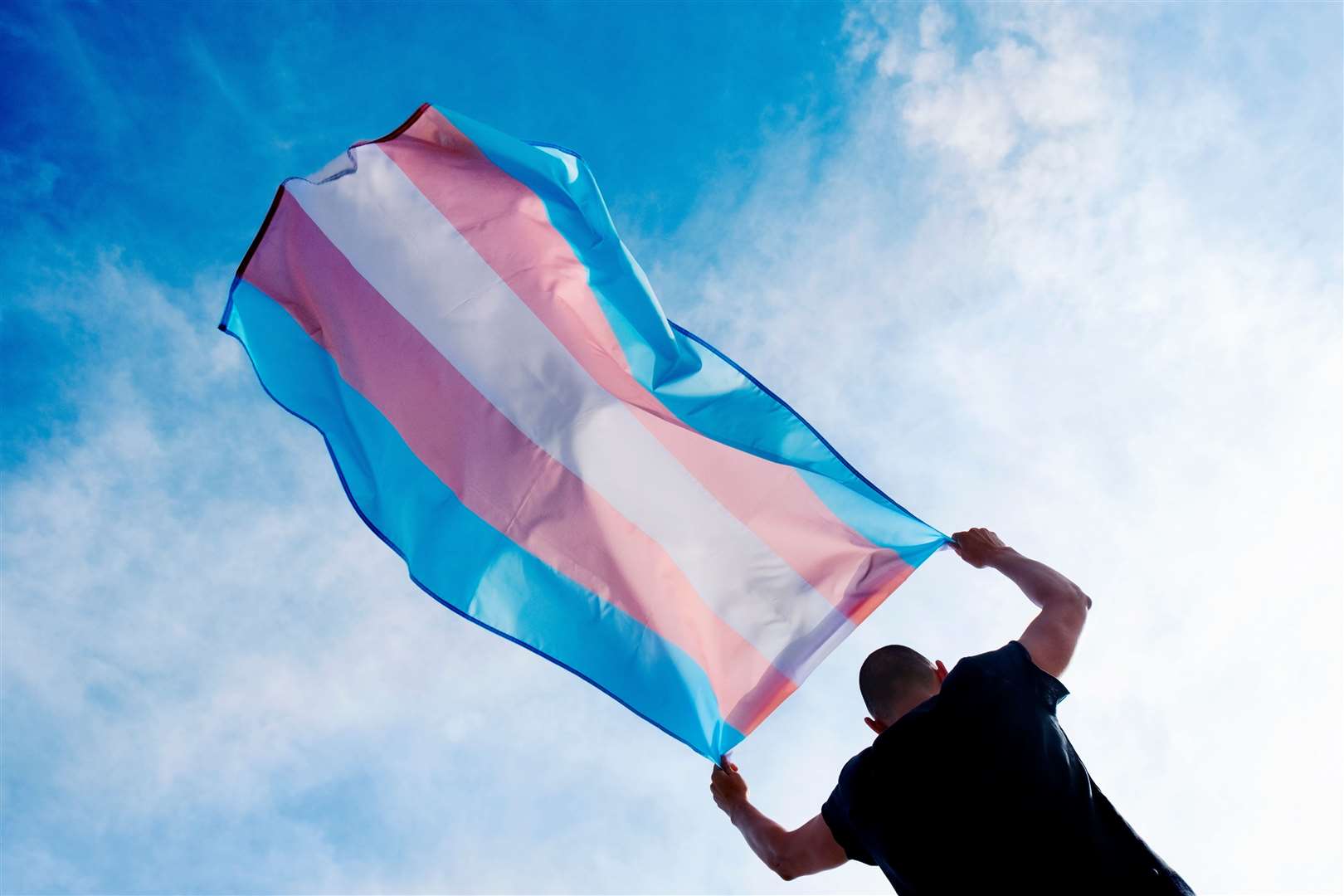 a young caucasian person, seen from behind, holding a transgender pride flag over his or her head against the blue sky