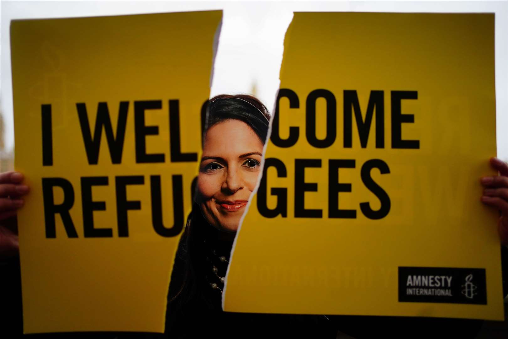 An Amnesty International campaigner wearing a Priti Patel mask tears up a ‘Refugees Welcome’ placard opposite the Houses of Parliament in London (Victoria Jones/PA)