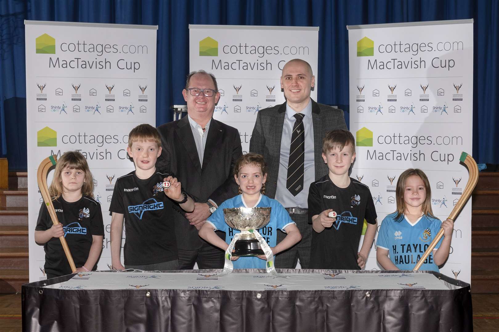 Camanachd Association chief executive Derek Keir and Shaun Gourley of title sponsors Cottages.com pose alongside Hilton Primary school kids who helped to conduct the first round draw. Picture: Neil Paterson