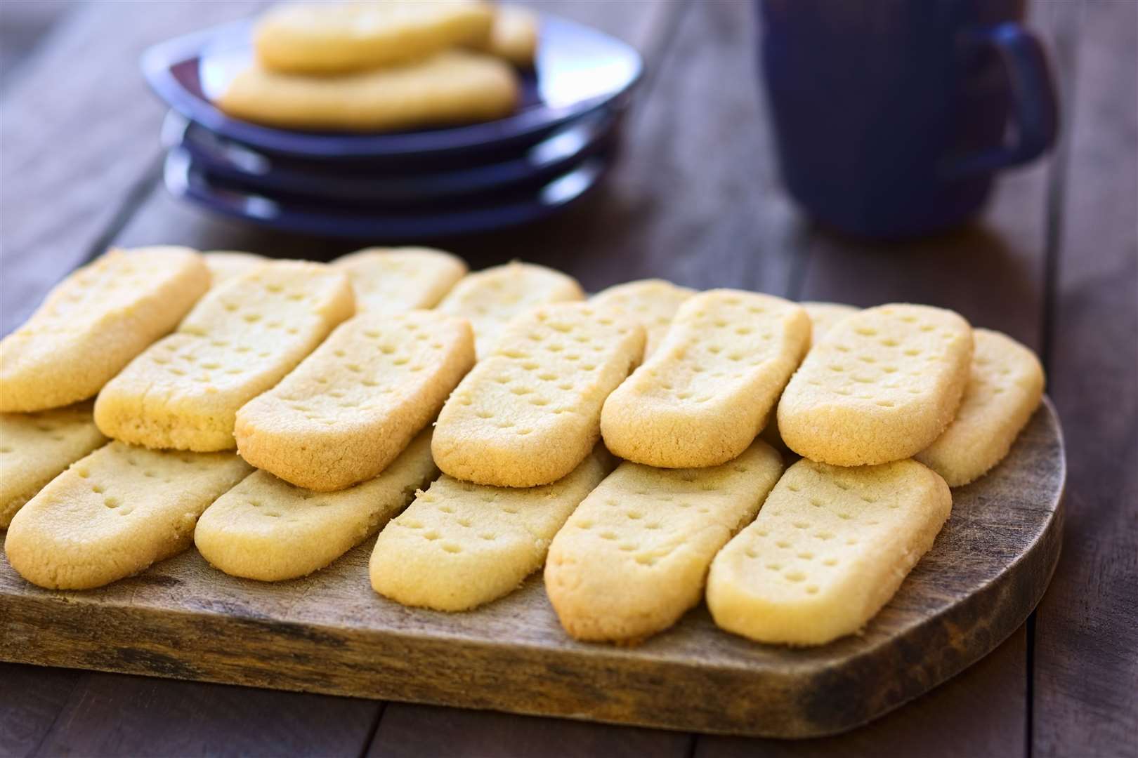 Homebaked shortbread biscuits on wooden board with plates and cup of tea in the back (Selective Focus, Focus on the front of the upper cookies)