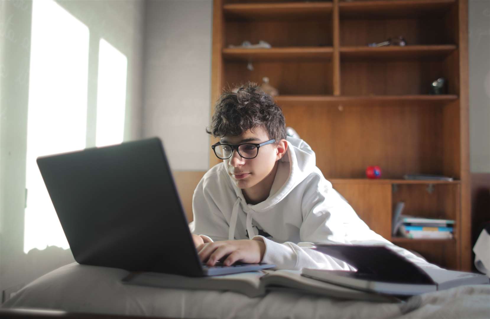 A young boy studies in his bedroom on books and laptop.