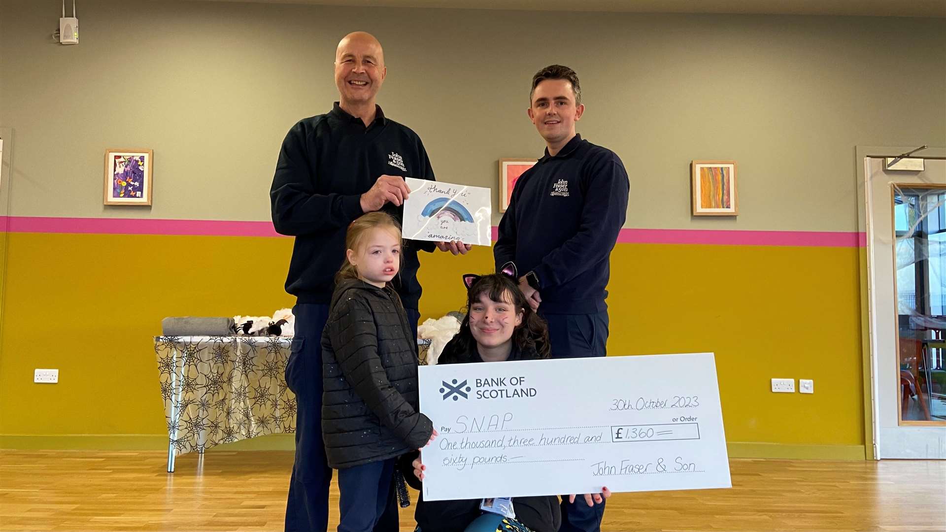 Memorial craftsman Drew Wheeler from John Fraser & Son (left) and trainee funeral director James Thompson, visit SNAP at The Haven Centre to hand over a fundraising cheque for £1360 to staff member Anya McPhee and one of the wee ones who was enjoying a play session in the charity’s new premises.